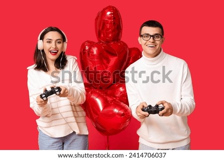 Young couple with heart-shaped air balloons playing video games on red background. Online dating