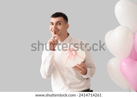 Handsome man with gift and balloons showing silence gesture on light background. Valentine's Day celebration