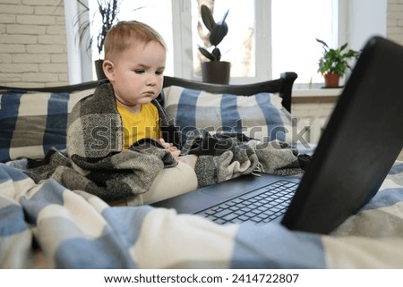 A handsome little boy sits on the bed, wrapped in a blanket, looking at the laptop screen. Sad boy in front of a laptop computer. Overall plan.