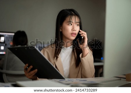A professional Asian businessman is talking on the phone with her supplier, having a serious phone call, working at her desk in the office. Royalty-Free Stock Photo #2414733289