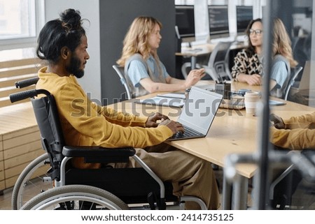 Young indian guy in wheelchair working on laptop sitting at office table with his coworkers