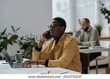 Black office worker talking on smartphone sitting at laptop, his colleague working on background