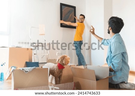 Gay couple decorating new apartment and putting up a framed piece of art on the wall in new home with their dog. Man in causal clothes hanging a painting on a wall.