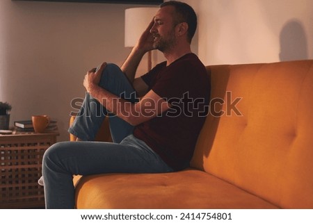Man with headache and knee pain sitting on a sofa at home. Royalty-Free Stock Photo #2414754801