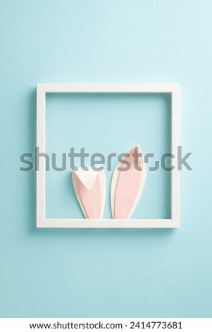 Playful Easter happiness scene. Overhead vertical shot of an enchanting Easter bunny hiding in a photo frame, ears popping out joyfully on a soft blue background