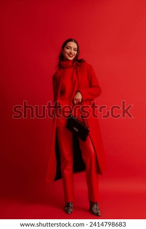 Fashionable happy smiling woman wearing trendy red midi coat, turtleneck sweater, leather pants, zebra print boots, holding baguette bag, posing on red background. Full-length studio fashion portrait Royalty-Free Stock Photo #2414798683
