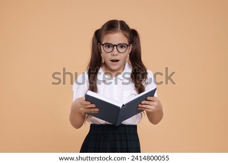 Surprised schoolgirl in glasses with book on beige background