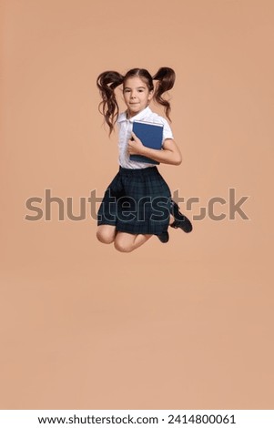 Cute schoolgirl holding book and jumping on beige background Royalty-Free Stock Photo #2414800061
