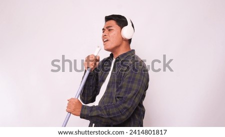 Asian man listening music with headphone and singing on broom against white background