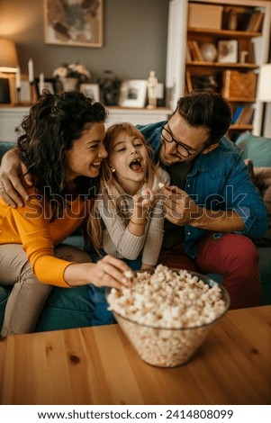 Dad, mom, and daughter sharing laughter on the living room sofa while watching a movie and enjoying popcorn