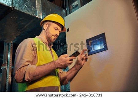 good looking pensive technician in safety clothes and helmet looking at his phone while working hard