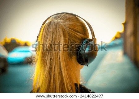 Close-up, rear view of a young girl's head with headphones, listening to music or audio. Long hair in a ponytail, set against an urban backdrop with soft sunset lighting.