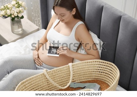 Beautiful pregnant woman with ultrasound picture of baby and basket on bed indoors