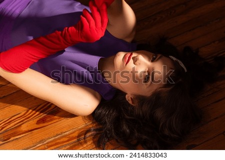 Artistic portrait with shadows: young caucasian model with black hair laying down on a wooden floor with half her face under the light, wearing Lila outfit and red long gloves. Perfect skin