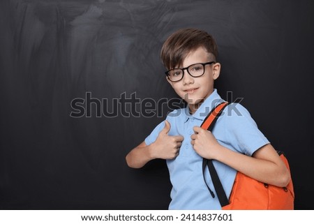 Cute schoolboy in glasses showing thumbs up near chalkboard, space for text