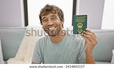 Cheerful young man, casually sitting on the living room sofa, joyfully holds his macao passport indoor, displaying a confidence-filled, happy smile at home.