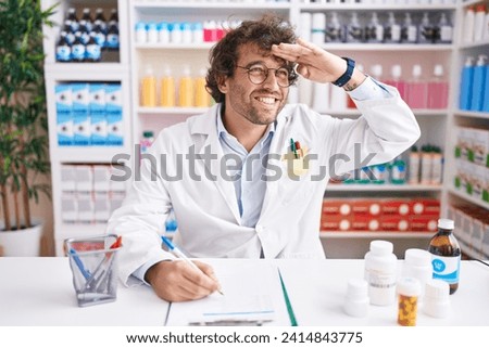 Hispanic young man working at pharmacy drugstore very happy and smiling looking far away with hand over head. searching concept. 