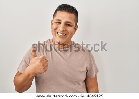 Hispanic young man standing over white background doing happy thumbs up gesture with hand. approving expression looking at the camera showing success. 