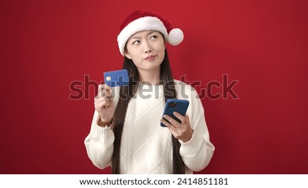 Young chinese woman wearing christmas hat using smartphone and credit card over isolated red background