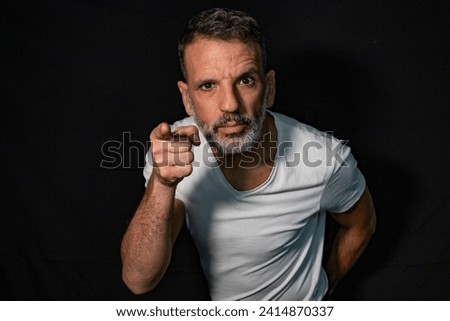 Portrait of a mature man with a gray beard, wearing braces, gesturing with his hands, wearing white t-shirt on a black background. Royalty-Free Stock Photo #2414870337