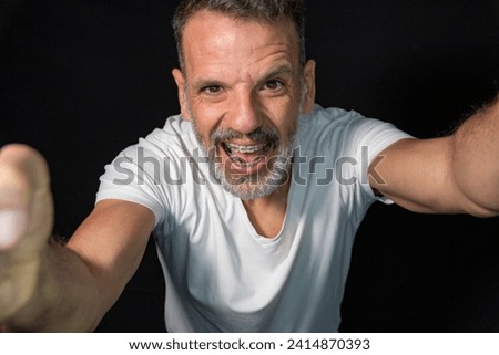 Portrait of a mature man with a gray beard, wearing braces, gesturing with his hands, wearing white t-shirt on a black background. Royalty-Free Stock Photo #2414870393