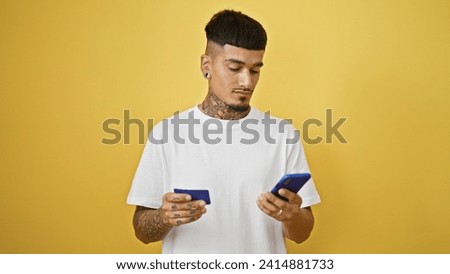Young, handsome latin man, tattooed and upset, haphazardly uses his credit card and smartphone against isolated yellow wall.