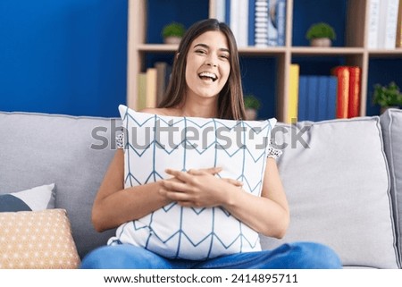 Hispanic woman hugging pillow sitting on the sofa smiling and laughing hard out loud because funny crazy joke. 