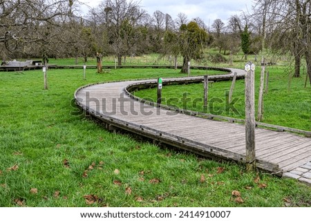 S-shaped winding wooden walkway bridge over green grass with hiking trail signs, bare fruit trees in Flanders organic orchard at Alden Biesen Castle, cloudy winter day in Bilzen, Limburg, Belgium Royalty-Free Stock Photo #2414910007