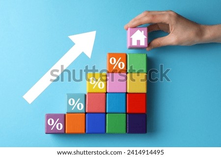 Mortgage rate rising illustrated by upward arrow. Woman putting cube with house icon near other ones on light blue background, top view