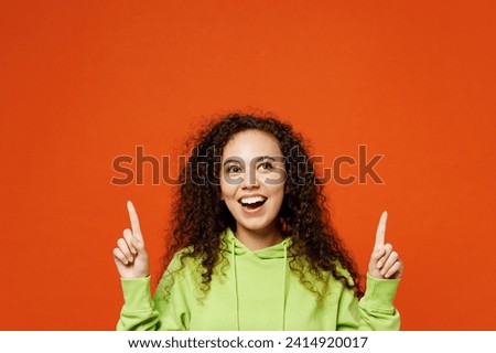Surprised fun amazed young woman of African American ethnicity wears green hoody casual clothes point index finger overhead on area mockup isolated on plain red orange background. Lifestyle concept