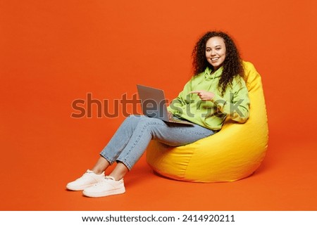 Full body smiling young IT woman of African American ethnicity wear green hoody casual clothes sit in bag chair hold use work point finger on laptop pc computer isolated on plain red orange background Royalty-Free Stock Photo #2414920211