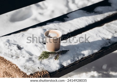 A paper cup of cappuccino coffee on a snow-covered bench in a winter park in sunny weather.