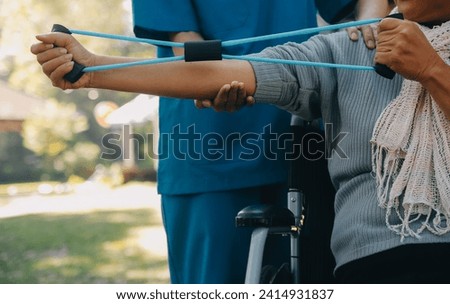 Young nurse or physiotherapist in scrubs helping a happy retired old woman do fitness exercises with light weight dumbbells at home. Concept of physiotherapy for seniors