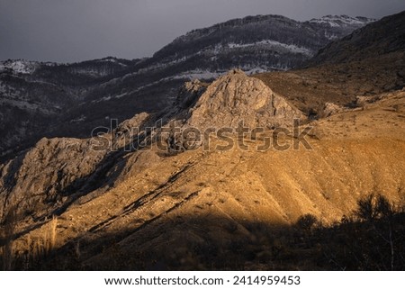 Landscape of rocks at classic golden hour glow, sunrise in snowy mountains, dramatic gray sky, snowy forest and yellow sun light, layers of mountains