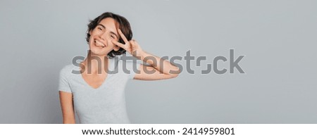Long banner image of young smiling happy cheerful friendly female with trendy hairstyle in casual clothes showing peace or victory gesture sign and looking on camera with funny grimace on face