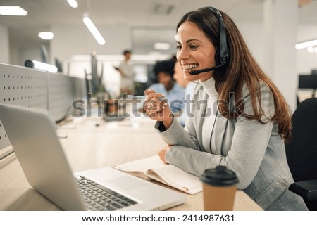Smiling caucasian business woman with headset working on a laptop in a call center on the line. Happy female working in a busy customer service department and taking notes while talking with a client. Royalty-Free Stock Photo #2414987631