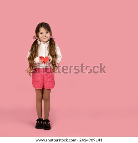 Stylish little girl in white shirt looking at camera Holding paper heart in hands, isolated by pink background, Valentine's day concept picture.