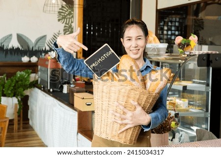 Asian young barista worker female holding Promotion sign board and bread in basket in front of bakery cabinet taking pictures to promote in social media channel, happy small business owner lifestyle