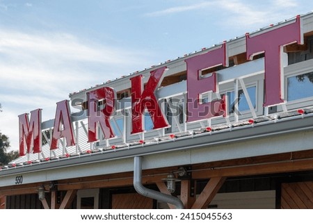 A vibrant red illuminated market sign on the roof of a rustic building. The storefront has a peaked roof, large window and wooden exterior wall. The background is blue sky with clouds and green trees.