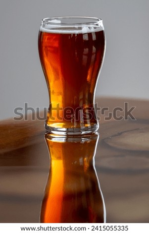 A frosted clear pint glass contains a red sour craft beer. The ale is on the corner of a wooden table. The top of the alcohol refreshment has white froth. The refreshing red color alcohol is a sample.