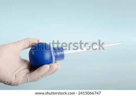 Bulb syringe, irrigation vacuum cleaner. Surgical instruments, isolated on light blue background, held by doctor's hand with white gloves. Closed angle. Copy space. Royalty-Free Stock Photo #2415066747