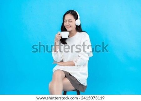 Asian female model with long hair Inhale the aroma and freshness of coffee while drinking and listening to music with gaming headphones. Sitting in a chair taking photos in a blue background studio