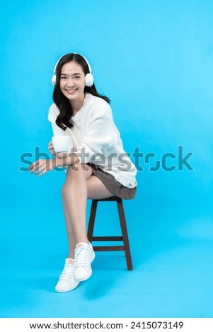 Asian female model with long hair Smiling happily listening to music with wireless headphones through the app and holding a cup of coffee. Sitting in a chair taking photos in a blue background studio