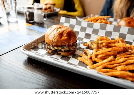 A delicious looking cheeseburger and french fries on on a tray with black and white checkered paper tray liner on a table in a fast food restaurant. American food. Hamburger with fries