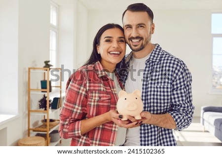 Joyful young couple stands in the living room at home, holding a piggy bank together. This portrait captures the essence of the importance of saving money and financial care within a family
