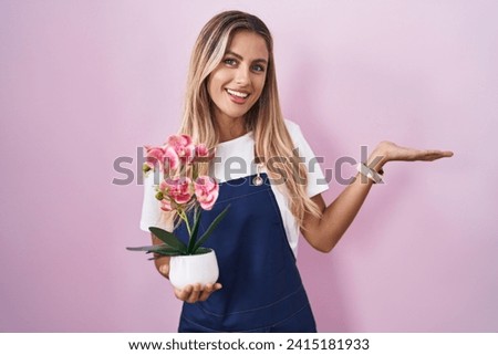 Young blonde woman wearing gardener apron holding plant smiling cheerful presenting and pointing with palm of hand looking at the camera. 