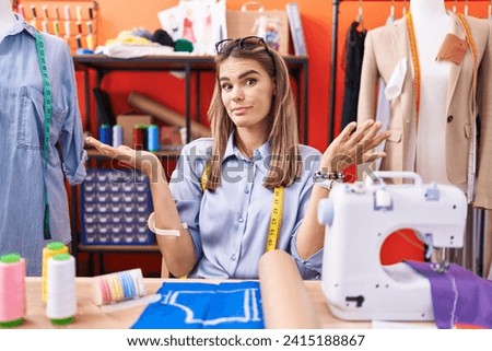 Hispanic young woman dressmaker designer at atelier room clueless and confused expression with arms and hands raised. doubt concept.  Royalty-Free Stock Photo #2415188867