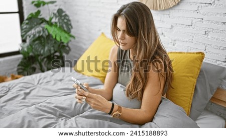 Young, beautiful hispanic woman sits relaxed on her comfortable bed, using her smartphone in the well-lit room of her apartment, depicting the everyday lifestyle of the modern adult. Royalty-Free Stock Photo #2415188893