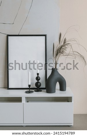 Empty frame mockup, modern black ceramic vase with dry grass, minimalistic candlestick on the dresser. Stylish home interior decor.