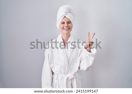 Blonde caucasian woman wearing bathrobe showing and pointing up with fingers number two while smiling confident and happy. 